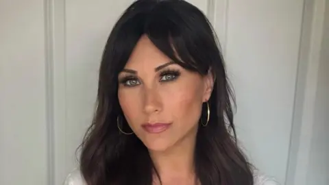 A close-up of Marina Berry-Dealey who has long dark brown hair across her shoulders. She is wearing large hooped silver earrings and is standing against a white panelled wall. Her white top can just be seen. 