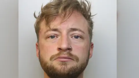 Avon and Somerset Police A police mug shot of a man with fair hairm blue eyes, a nose ring and a beard looking directly at the camera.