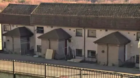 A row of RAAC affected properties. Each house has four windows, some are open. The buildings are grey concrete. 