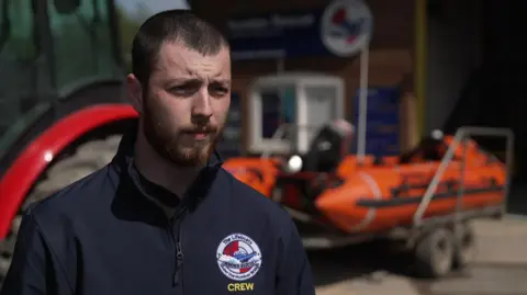 A man with dark hair cropped short and a dark beard. He wears a dark jacket with the Humber Rescue logo on it and stands in front of one of the vessels.