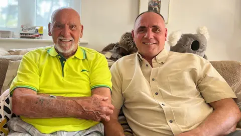 Jim and Sean are sitting on a sofa in a living room, smiling toward the camera, with patterned cushions, a window behind, and framed photos on the wall.