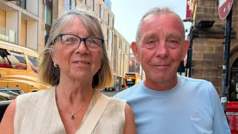 BBC/Adam Samuel Brenda and Eddie smiling into the camera. Brenda is on the left and has short blonde hair and is wearing dark framed glasses with a beige striped top on. Martin has grey short hair and is wearing a blue t-shirt. They are standing on a busy street with buses going past.