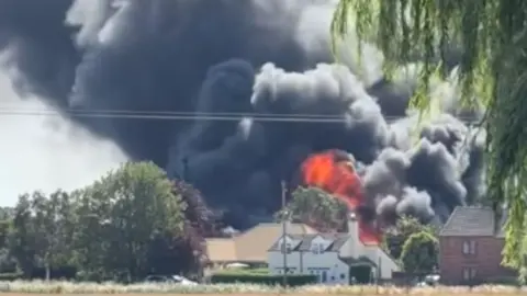 Large black smoke and orange flames are billowing into the sky from behind a building. In front of the building is a large open field with trees either side.