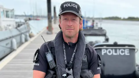 Stuart Woodward/BBC A man wearing a police uniform and black life jacket looks directly at the camera with a stern look on his face. He is standing on decking in front of a harbour and a police boat. 