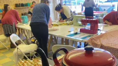 Spark Somerset A group of people stood around a table with sewing machines on it with a pot of tea and a cake in the foreground