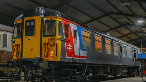 James Cole/Clacton Express Preservation Group The Clacton Express electric train is pictured in a storage unit with its front having been given fresh paint in yellow and some red, white and blue. The rest of the train's body is grey.