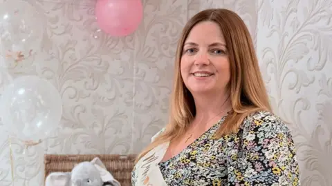Supplied A pregnant woman at a baby show with a floral dress on. A framed picture and a hamper with toys is in the background