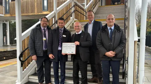 Five men in suits stand at the foot of the stairs of the new bridge. The transport minister Peter Hendy holds a plaque.