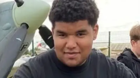 A young man with dark curly hair smiles and wears a black t-shirt. 