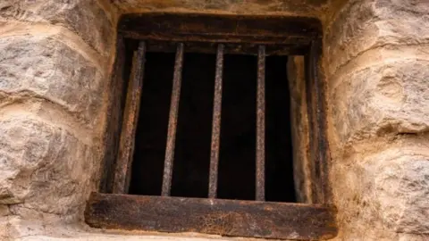 David Brough A close up of a metal-barred opening of the stone structure. The bars are rusty brown. 