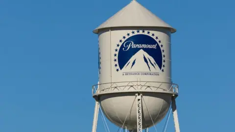 A white water tower with the dark blue paramount logo of a mountain inside a circle of starts sits against a bright blue sky.