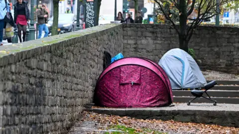 A pink and a blue tent are pitched on concrete steps. On the other side of the wall people walk past carrying shopping bags.