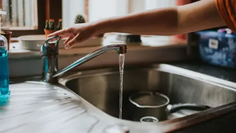 A person's right hand reaches towards a tap at a kitchen sink, the sink it stainless stell, the tap is running and there is a saucepan in the sink. The person is about to turn off the tap, which is running with water and there is a windowsill with plants in the background.