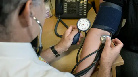 An over the shoulder view of a man using a stethoscope and a blood pressure machine on the arm of another person who is sat in front of him