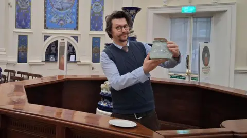 Dr Jed Atkinson A man with a curled mousache holds a jar of water as he stands in a museum in Harrogate 