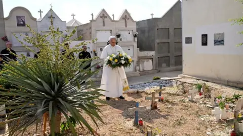 Reuters The Pope holds is holding flowers at a cemetery. Behind him are officials dressed in black suits and a row of white buildings with crosses on them.