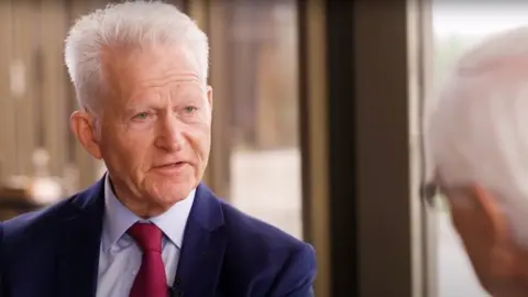 IsHLT Usa ka tigulang nga lalaki nga adunay hait nga puti nga buhok, nga nagsul-ob sa usa ka navy blue suit, light blue shirt ug mapula nga ihigot, nga giinterbyu sa usa ka lalaki nga adunay bukobuko sa.