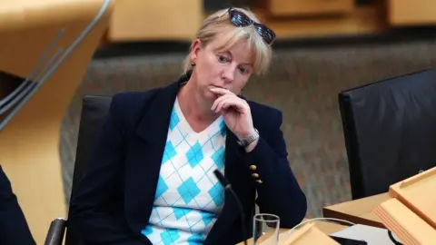 PA Media Shona Robison - a woman wearing a dark jacket and blue and white top - sits frowning in Holyrood. She has a pair of sunglasses on top of her head.
