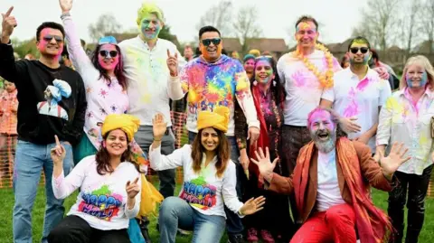 Amith Nair A group of men and women celebrating Holi posing for a picture. They have a variety of bright colours such as pink and green dispersed on them. Behind an orange mesh barrier other people can be seen gathering.