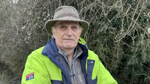 BBC/ Kate Bradbrook Rob Newby stands in front of a bramble. He is wearing a neon high-visibility jacket and tweed hat. 