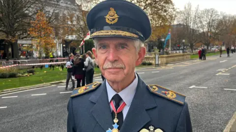 An elderly man dressed in military uniform. He has light grey/white hair and a moustache. He is looking at the camera.