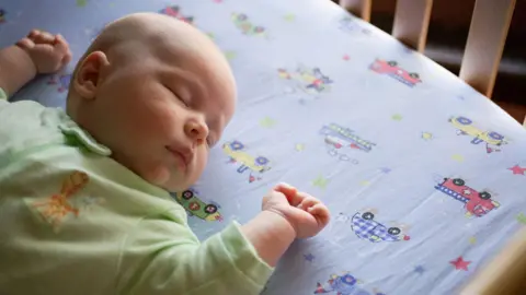 An overhead shot of a sleeping baby wearing a green onesie lying on a bedsheet with cars and trucks.