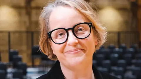 PA Media A woman with short blonde hair and black rimmed glasses, wearing a black top in a room with rows of black seats and a brown wall