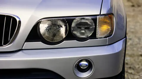 The front of a silver car with the headlights on one side visible