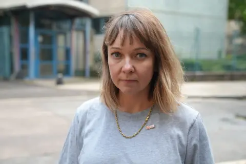 A woman stands outdoors on a city street, facing the camera. The person has shoulder-length light brown hair with short fringe and wears a grey T-shirt with a gold chain necklace. A blurred building entrance with blue framing and an urban street scene appear in the background.