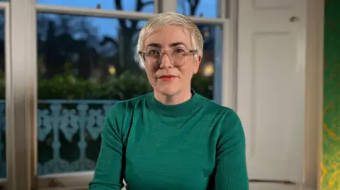 Branwen Jeffreys / BBC Alice Sullivan sits in a room looking at the camera. She has short platinum blonde hair, glasses and bright lipstick. She's wearing an emerald green jumper. 