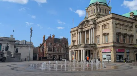The fountain in Queen Victoria Square, Hull, is located in front of City Hall. A seating area is visible in the background.