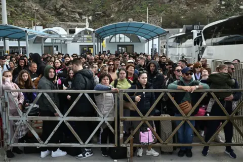 Getty stands behind the gates with a large group of people in a mix of ages carrying bags. They stand in front of the ferry terminal and there is a large bus next to them