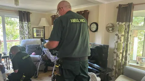 Two men in green South Western Ambulance Service uniforms treating a poorly woman in her front room.
