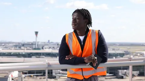Jake Mobbs/PA David Larbi is standing at a high viewpoint with the airport's control tower in the background behind him. He is wearing a hi-vis orange jacket over a dark zip up top, with a white T-shirt underneath and is holding a pen and notebook as he looks into the distance.