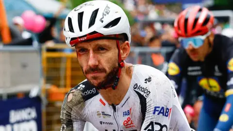 Adam Yates in a torn cycling kit and helmet, both predominantly white