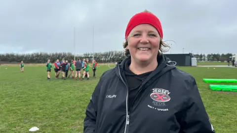 Julia is wearing a bright red beanie. She has short blonde hair underneath and is wearing a black jacket with the Jersey Reds rugby club logo on it.