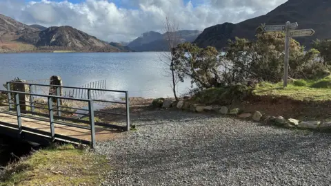 LDNPA A bridge with metal handles crosses a stream which opens up into a large lake beyond which mountains rise. A wooden finger post points in three directions on the edge of a gravelly path 