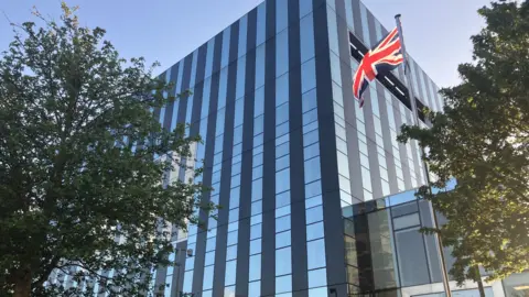 Nadia Lincoln/LDRS A union flag on a flag pole outside a large glass cube building. there are two trees one on each side of the buidling.
