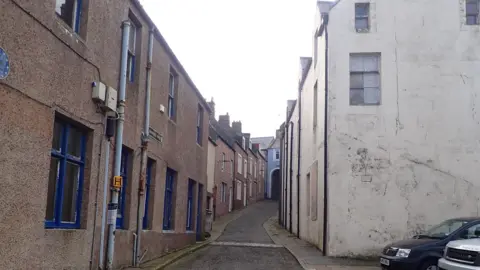 A narrow lane with buildings up both sides and a cobbled pathway in between