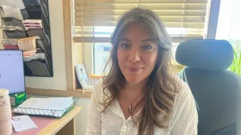 BBC Dr Jessica Langtree-Marsh is sat in her clinic. She is wearing a white shirt and has brown hair. She is sat next to a computer. 