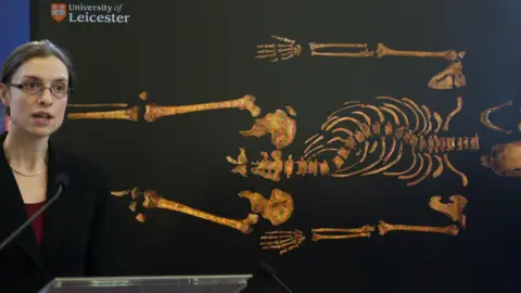 Getty Images A woman stands at a podium giving a speech in front of a picture of some bones.