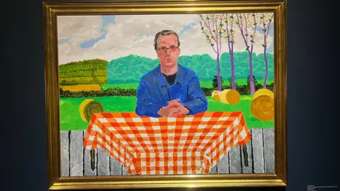 A vibrant painting showing a man in a blue jacket with brown swept back hair and round glasses who is sat at a table with an orange table cloth outside. 