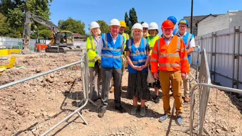 The image shows a group of men and women wearing hi-vis jackets and hard hats, standing on a construction site. It is a sunny day. The people are standing shoulder-to-shoulder and smiling at the camera. Metal barriers are separating the group from the site. Machinery is in the background.
