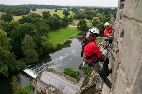 PA Two people in red tshirts and white helmets are abseiling stone castle walls with a view of trees and fields in the background