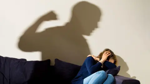 A reconstruction has been choreographed for a photo depicting an example of domestic abuse. A woman is cowering on a sofa protecting her head. The shadow of a man is cast on the wall, appearing to raise his fist ready to punch.