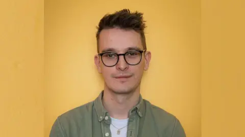 Youth Moves Matt Justice standing against a yellow wall. He is wearing a green shirt, white t-shirt and black framed round glasses. He has short brown hair which sticks up and is smiling faintly at the camera. 