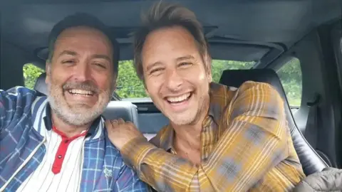 Vic Irvine and Chesney Hawkes are sat in the front seats of a car smiling at the camera. Mr Irvine sits to the left and has black hair flecked with greys and a grey beard. He wears a white polo top with black stripes and red detailing on the button strip and a blue tartan print bomber jacket over the top. Chesney Hawkes has his hand on Mr Irvine's shoulder, he has blonde short hair and wears a mustard yellow tartan print shirt.