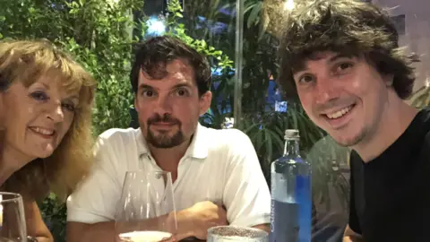 Family handout Oliver Robinson (right), Alexander Robinson (middle) and their mother (left) sit at a table at what appears to be a restaurant. There is decorative foliage in the background and they have wine glasses and a bottle of water on the table. Oliver is wearing a black tshirt, medium length brown hair, Alexander is wearing a white shirt with short dark hair and only their mum's face can be seen, she is smiling with longer strawberry-blonde hair.