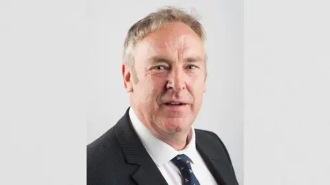 Gloucestershire County Council Headshot of councillor Vernon Smith who is wearing a dark blazer and tie and has short light brown/grey hair