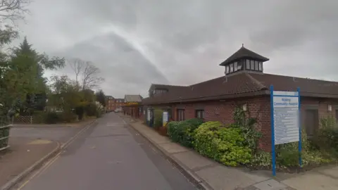 Google A view of the outside of Woking Community Hospital showing the main road leading to the building, along with signage.